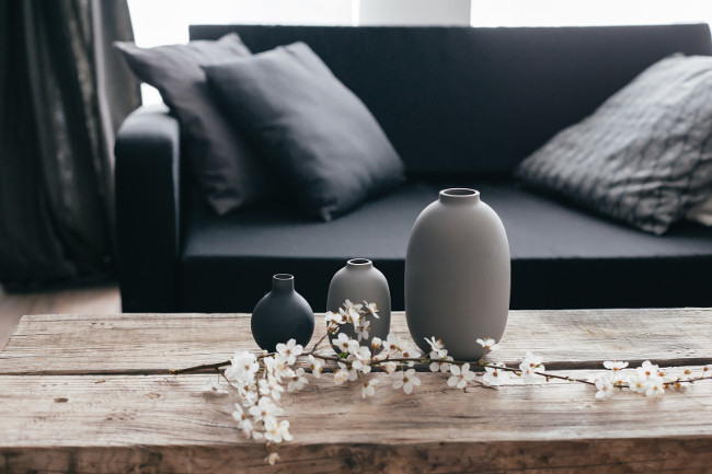 Minimalistic home decor on rustic coffee table over black sofa with cushions. Grey vases and spring flowers on wooden bench in small dark room interior. Scandinavian home style.