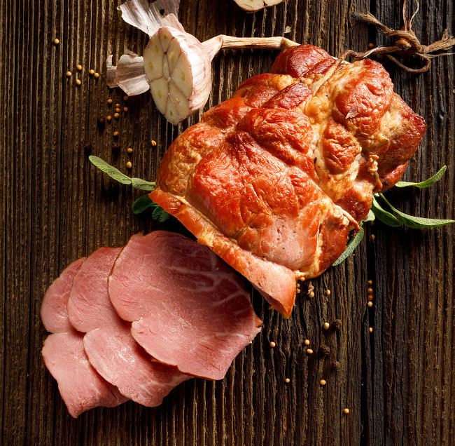 Sliced smoked gammon on a wooden  table with addition of fresh  herbs and aromatic spices, top view. Natural product from organic farm, produced by traditional methods