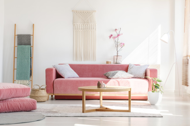 Soft, warm living room interior with a cozy, velvet sofa, millennial pink decorations and a beige macrame on a white wall