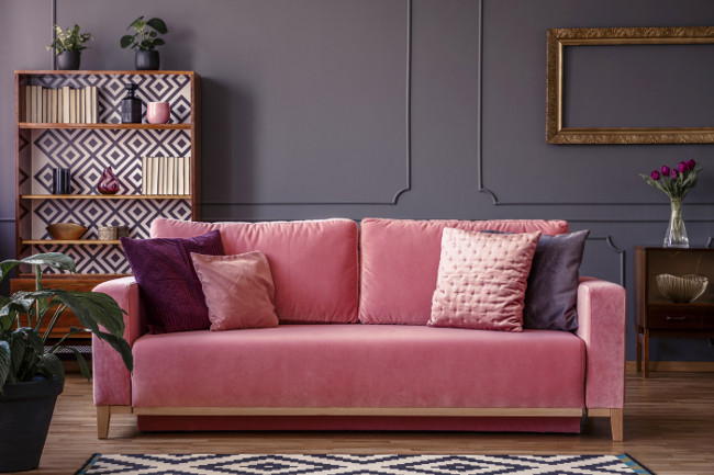 Cushions and blankets on a powder pink velvet sofa in a luxurious gray living room interior with wooden furniture