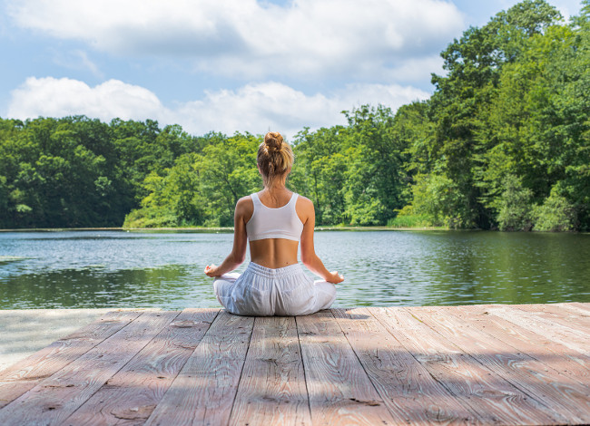 Attractive woman is practicing yoga and meditation sitting in lotus pose near lake. Young woman is meditating outdoors in morning.