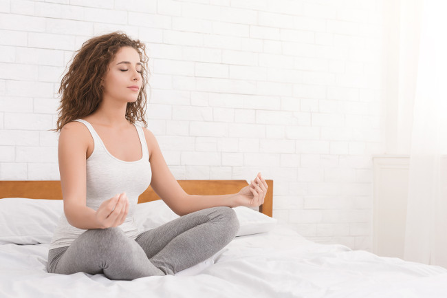 Meditation. Young millennial woman meditating in bed, sitting in lotus position on bed. Girl practicing anti-stress yoga, copy space