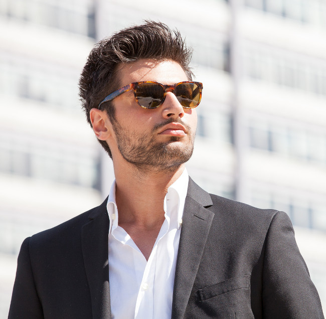Sexy gorgeous stylish man. Sunglasses. City style. A beautiful and charming man with sunglasses outdoors. Stubble and blacks hair. Intense light. White shirt and stylish jacket.
