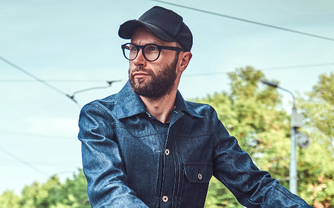 Attractive trendy man posing next to his bicycle. He is holding his bicycle. Man is wearing denim, cap and glasses. He is looking away. Street at the background.