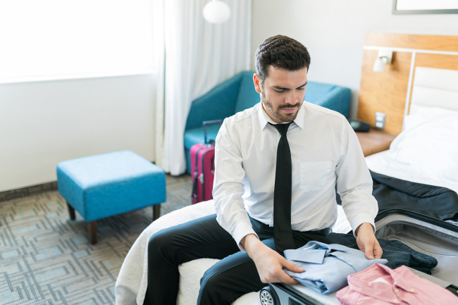 Mid adult Latin businessman packing clothes in suitcase while sitting on bed in hotel room