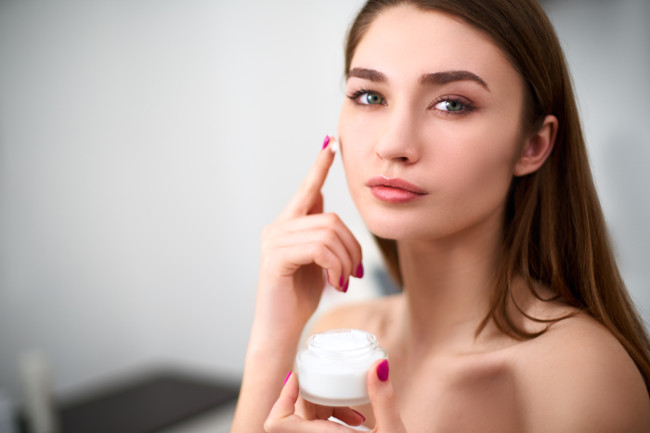 Smiling young mixed race woman applying face cream on her perfect skin. Model with flawless skin holding white jar of cream on blurred grey background in cosmetology clinic.
