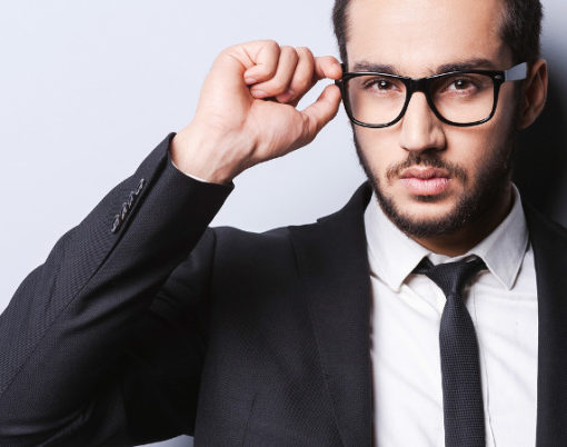 Portrait of handsome young man in formalwear adjusting his glasses while standing against grey background