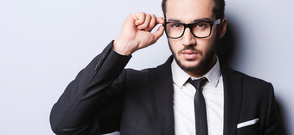Portrait of handsome young man in formalwear adjusting his glasses while standing against grey background