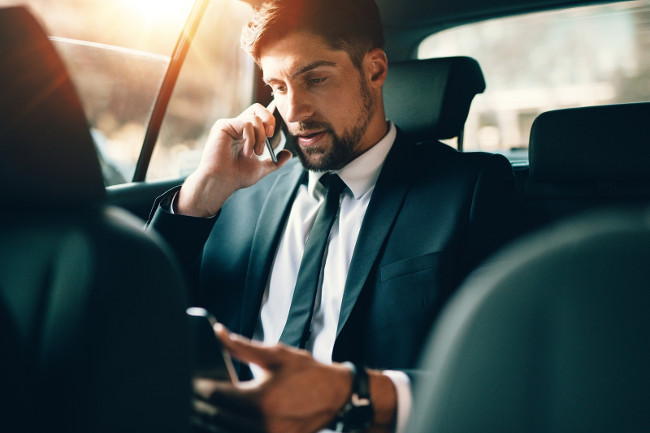 Young businessman talking on mobile phone and using tablet pc while sitting on back seat of a car. Caucasian male business executive travelling by a taxi and looking at digital tablet.