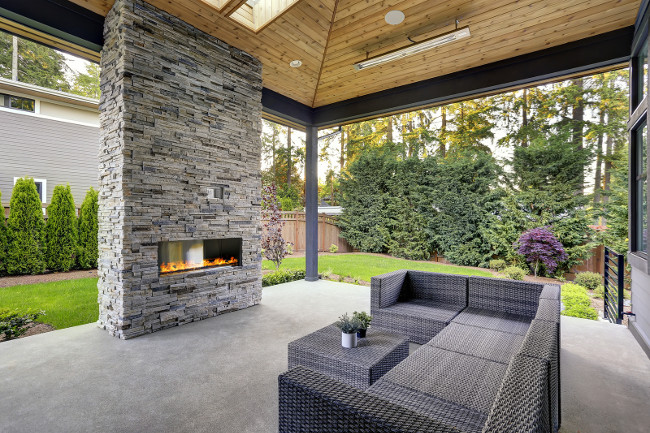 New modern home features a backyard with covered patio accented with stone fireplace vaulted ceiling with skylights and furnished with gray wicker sofa placed on concrete floor. Northwest USA