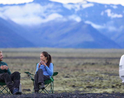 Travel couple by mobile motor home RV campervan. People sitting in chairs relaxing camping and enjoying traveling on Iceland in recreational vehicle. Young couple enjoying coffee in nature landscape.