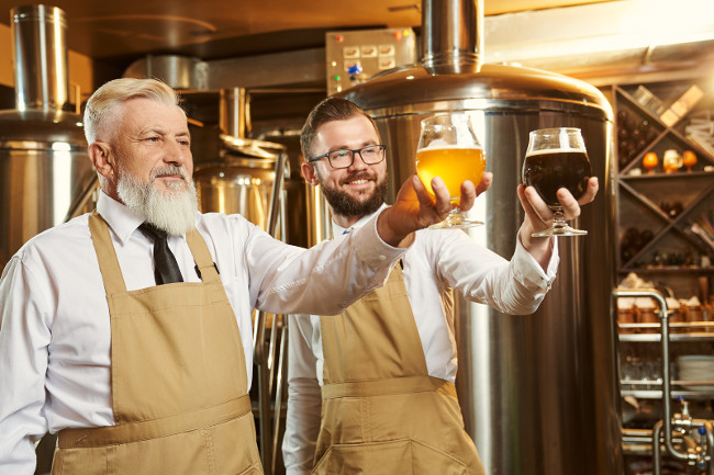 Two specialists standing with glasses of beer, looking at glasses and smiling. Brewery workers in white shirts and brown aprons posing at brewery background. Concept of craft beer brewery.