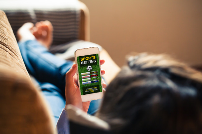 Woman holding a mobile phone with sports betting website app in the screen, while lying down at home at home.