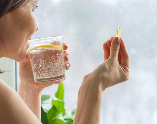 Woman takes pill with omega-3 and holding a glass of fresh water with lemon. Picture of the house, in the morning near the window. Vitamin D, E, a fish oil capsules. Nutrition, healthy eating, lifestyle