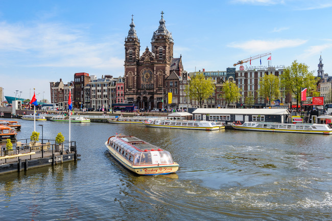 Amsterdam, Netherlands - April 18, 2019: Tourists sightseeng at Canal Boats near the Central Station of Amsterdam. View to Basilica of Saint Nicholas