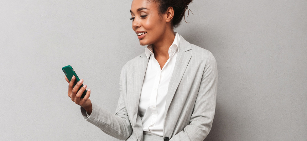 Full length portrait of a confident african business woman wearing suit sitting on a chair over gray background, holding takeaway coffee, using mobile phone