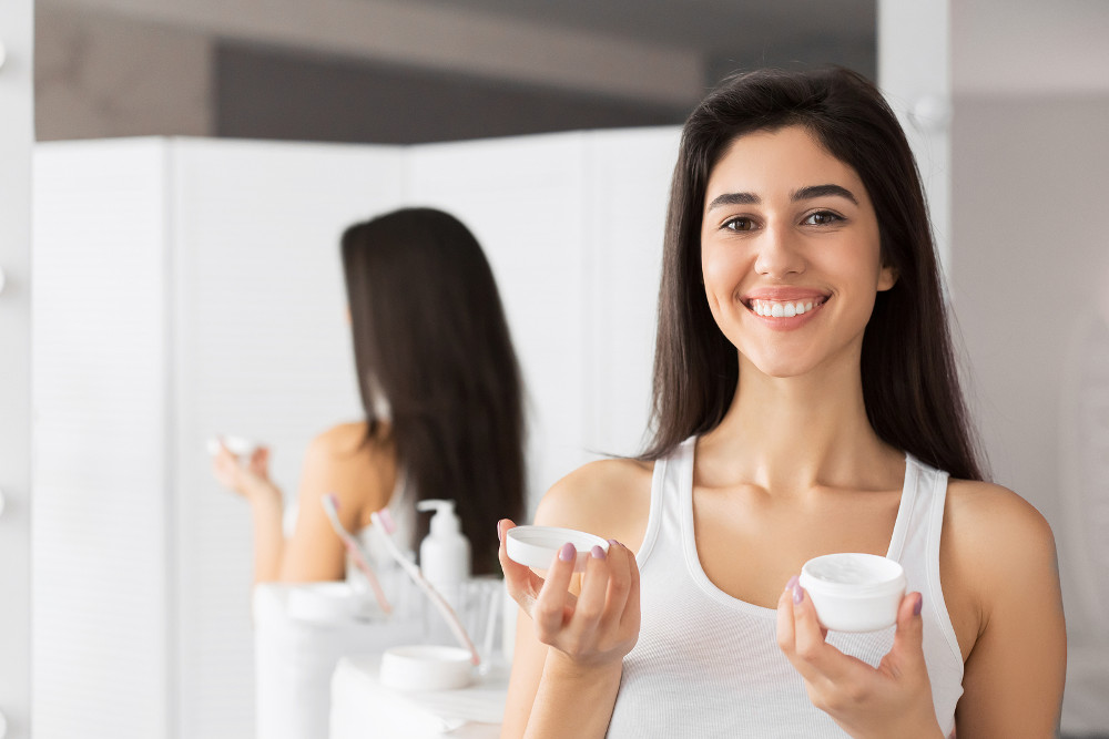 Happy Young Woman Holding Face Cream Cosmetic Product Standing In Bathroom. Daily Skincare Routine. Selective Focus