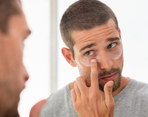 Young man taking care of his undereye wrinkles putting anti aging eye moisturizer. Handsome guy applying moisturizer and looking at himself while standing in front of the mirror in the bathroom.