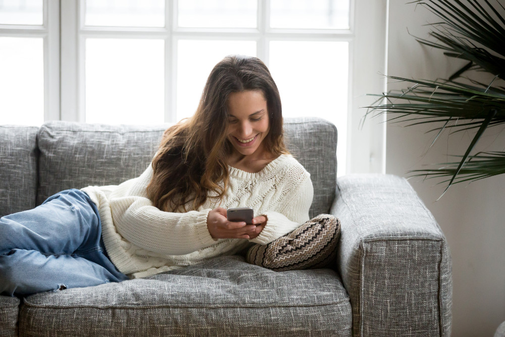 Happy Young Woman Lying On Sofa At Home Using Smartphone