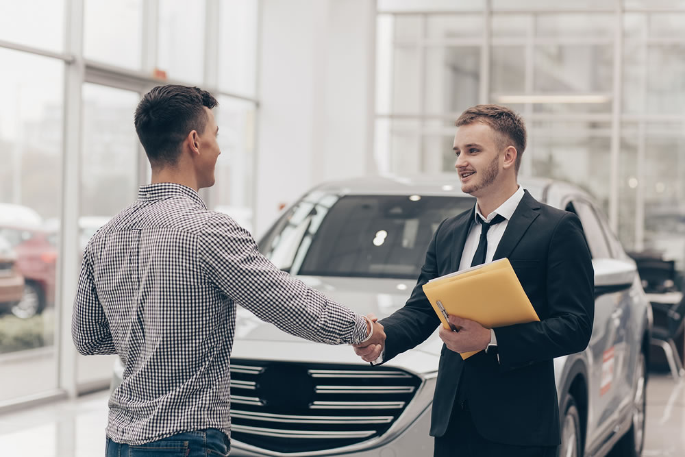 Professional car salesman smiling, shaking hands with his male customer in front of a new automobile