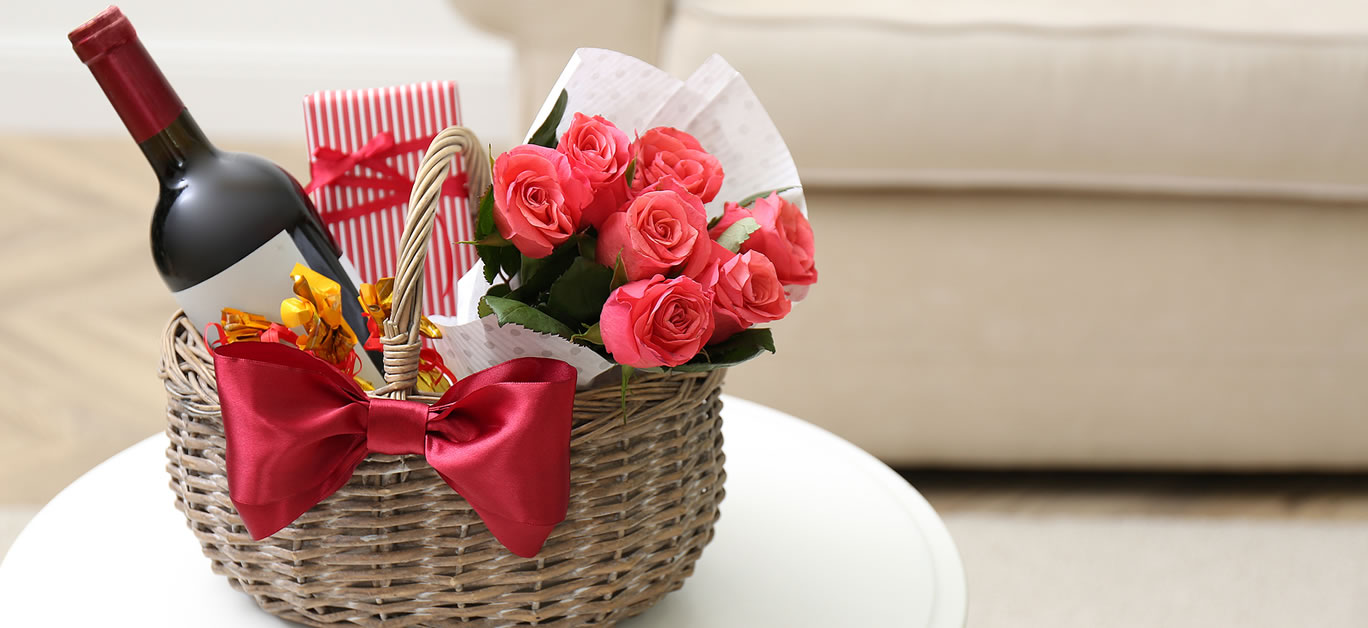 Wicker basket with gift, bouquet and wine on white table indoors.
