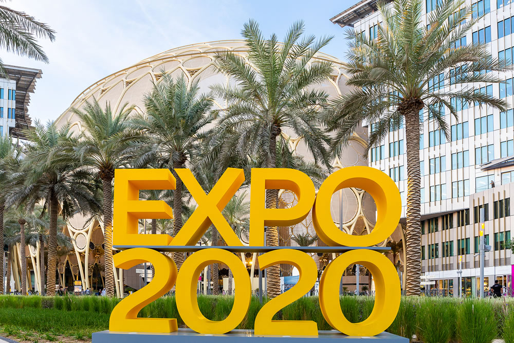 Expo 2020 yellow logo sign at Expo 2020 Dubai with Al Wasl Plaza and palm trees in the background.