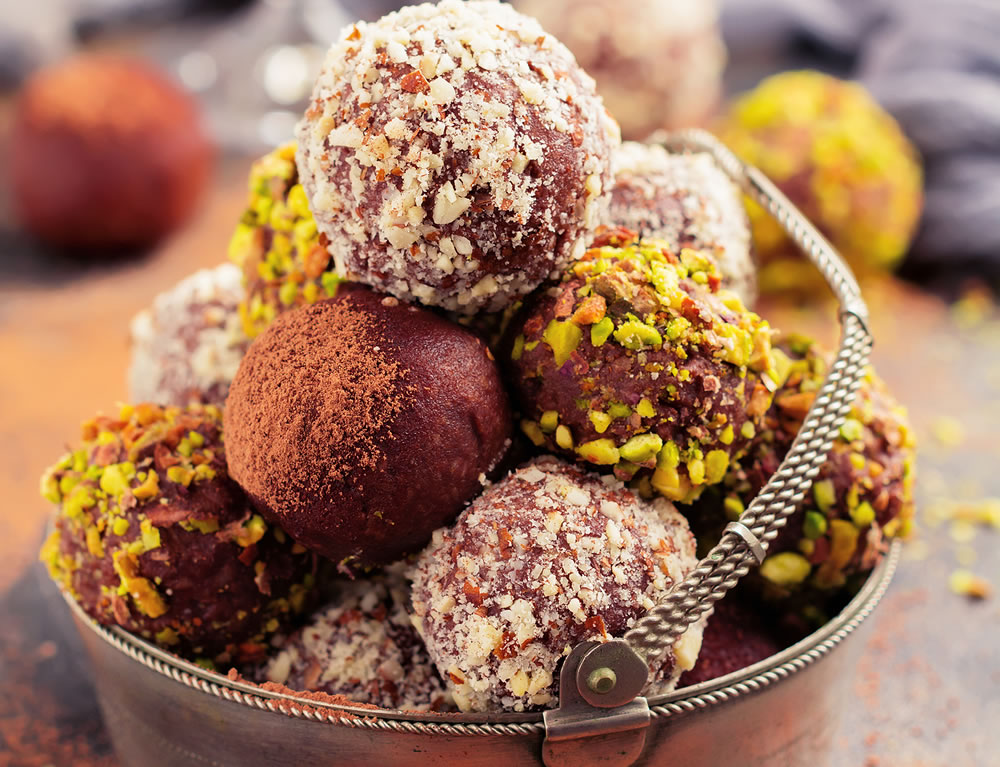 Variety of homemade dark chocolate truffles with cocoa powder, pistachios, almonds in dark brown background texture