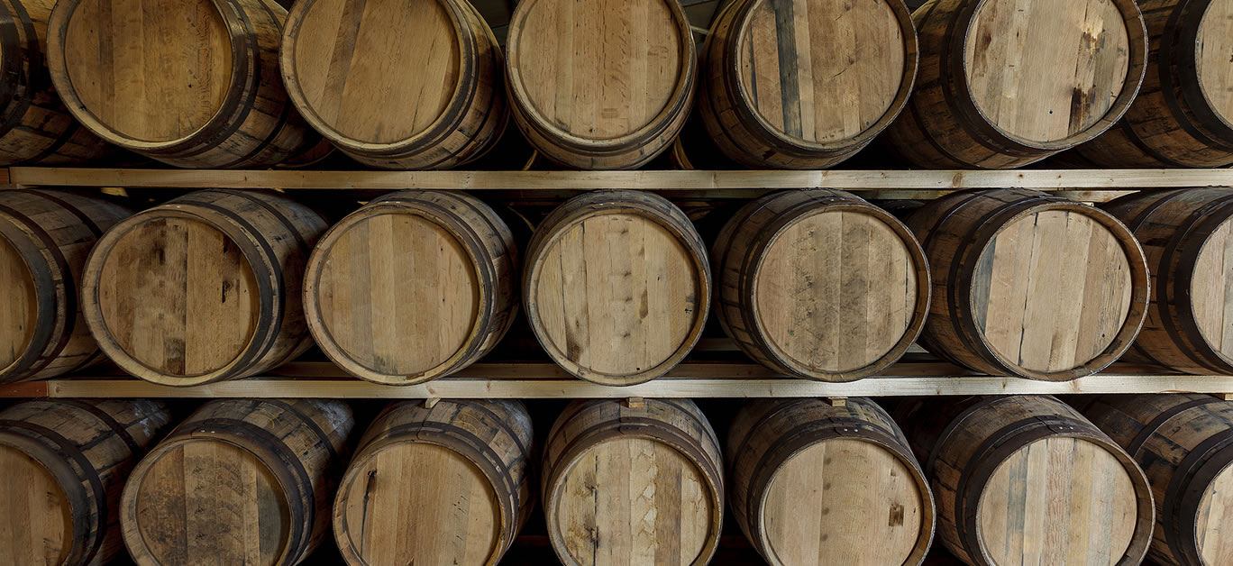 A front on view of a row of stacks of traditional full whisky barrels, set down to mature, in a large warehouse facility