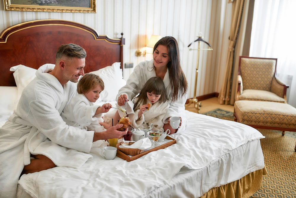 Parents and two kids in white bathrobes smiling while having breakfast in bed, eating and drinking in luxurious hotel room