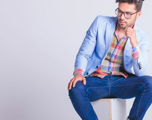smart casual man seated on box, wearing glasses and jeans, posing while looking away in studio background