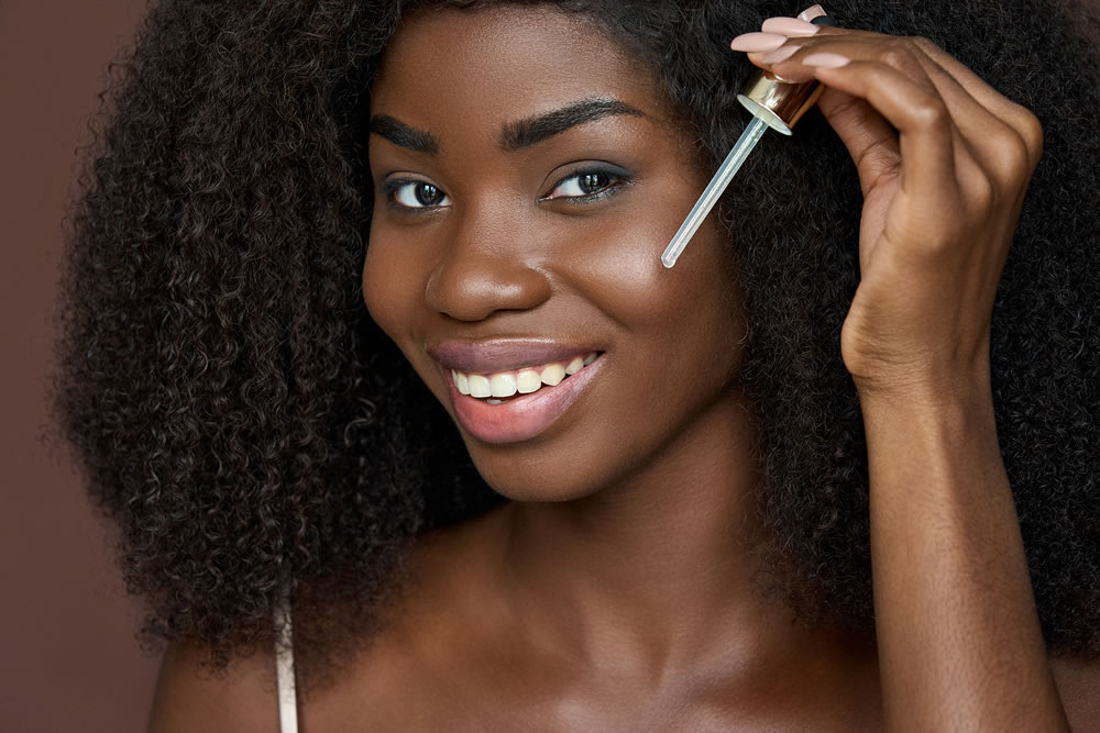 Smiling happy beautiful young African black woman using dropper applying hyaluronic serum