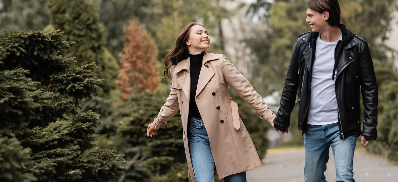 cheerful and stylish couple in autumnal outfits holding hands while walking in park.