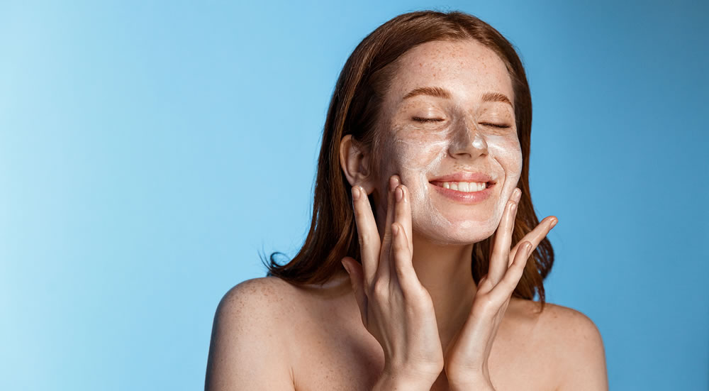 Smiling redhead girl with freckles washing her face, using cleanser, skin care gel after shower, standing over blue background