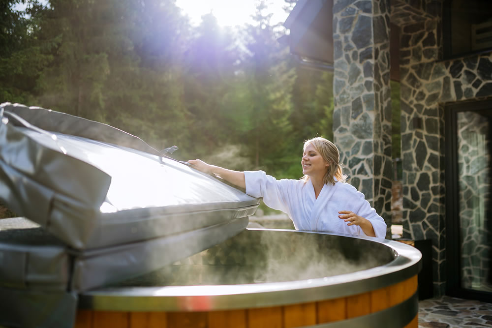 Woman in bathrobe opening lid of hot tub, checking temperature, ready for home spa procedure in hot tub outdoors