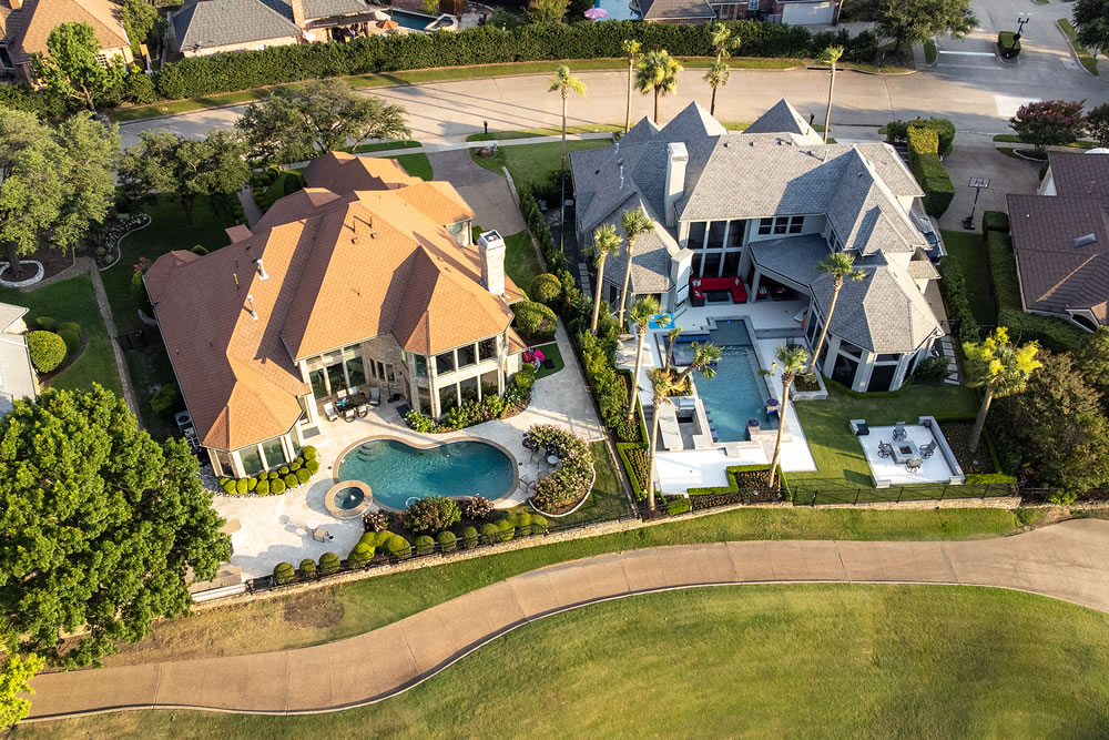 Aerial drone view of luxury mansions with swimming pools surrounded by green grass and trees in the summertime