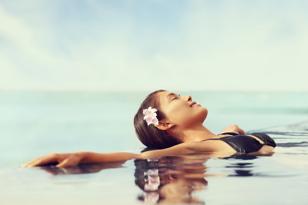Luxury resort woman relaxing in infinity swim pool. Asian young adult lying down in swimming pool of beach resort for summer holidays or travel vacations.