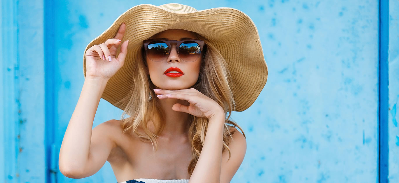 Woman portrait on blue background, sitting on the steps in a nice big straw hat and sun glasses, red lipstick and beautiful white teeth