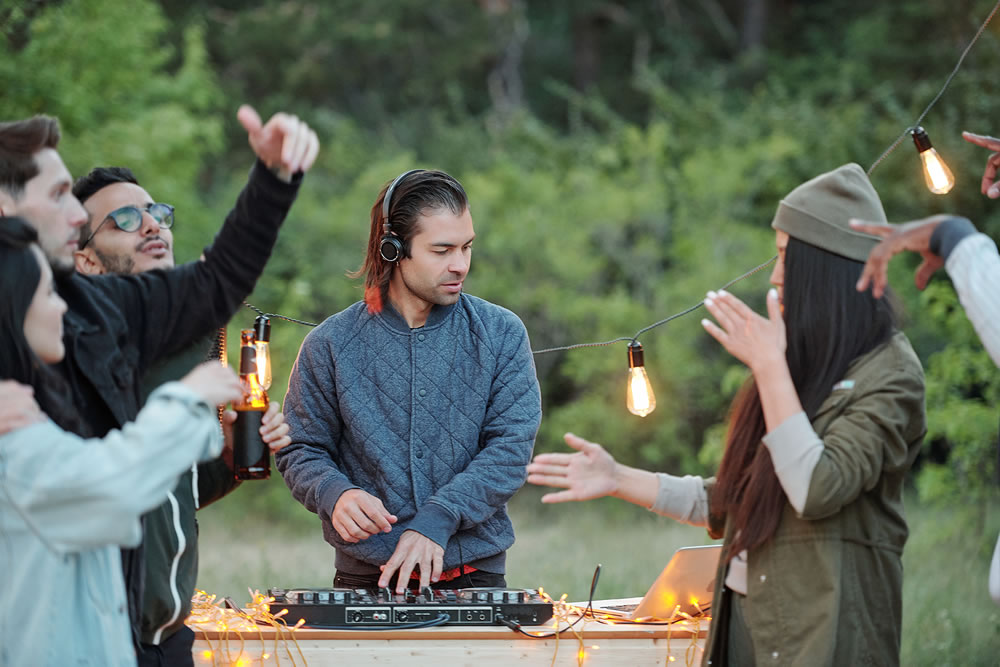Young casual man in jacket and headphones making music for his joyful intercultural friends dancing and enjoying summer weekend