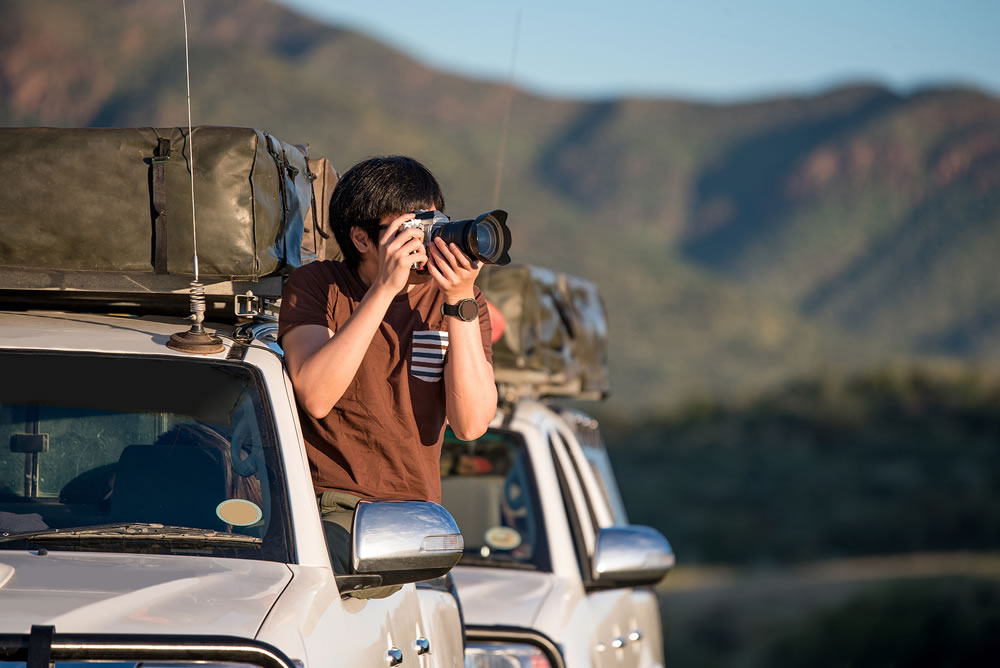 Young Asian man traveler and photographer sitting on the car window photographing sunrise during road trip in Namibia, Africa. Travel photography concept