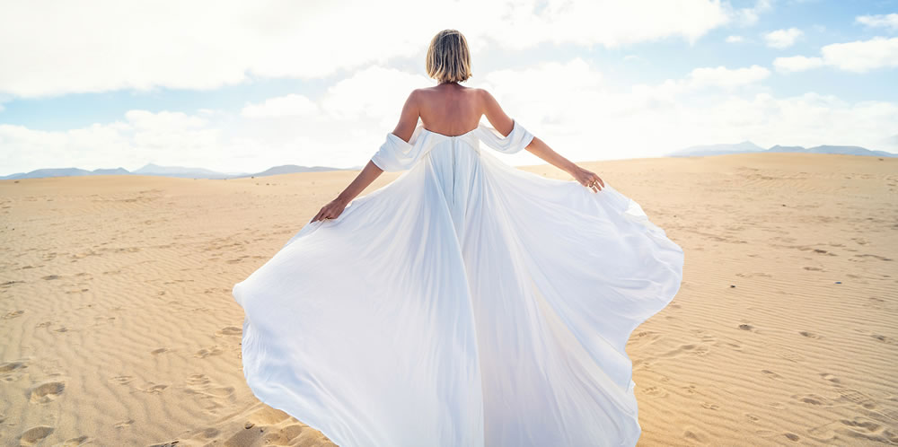 Back view of a blonde woman in fashionable, maxi white wedding dress posing on the desert, dancing in sunny light