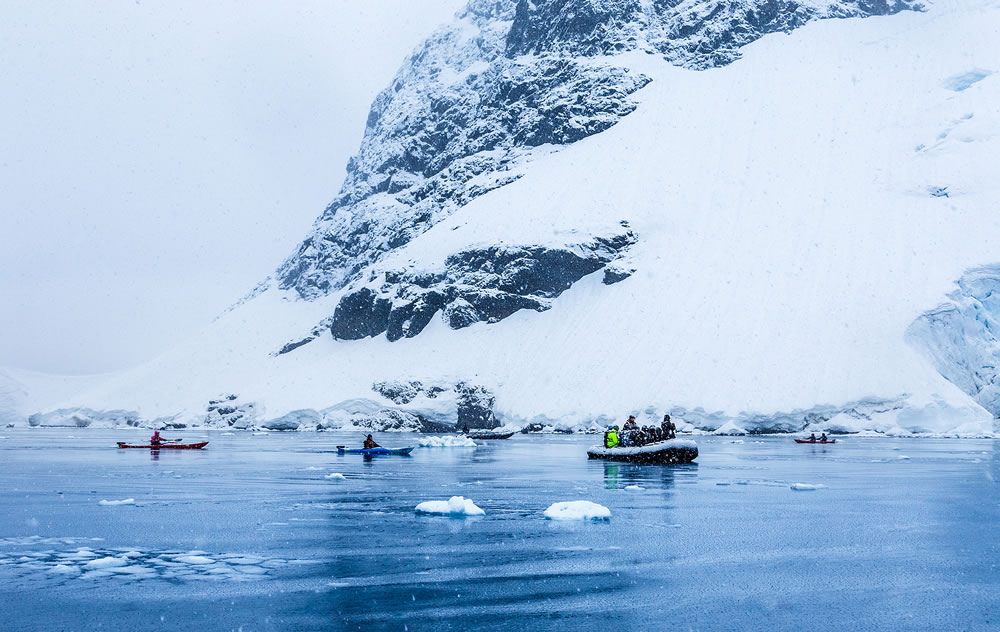 Snowfall over the motor boat with tourists and kayaks in the bay with rock and glacier in the background, near Almirante Brown, Antarctic peninsula