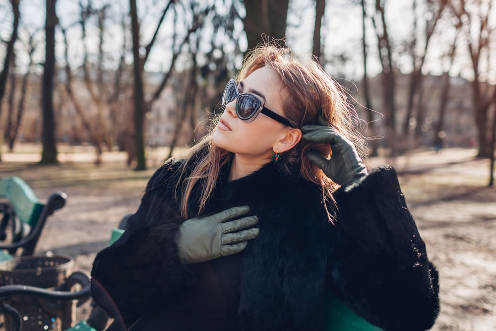 Close up portrait of stylish young woman wearing fur coat, green leather gloves and sunglasses in park