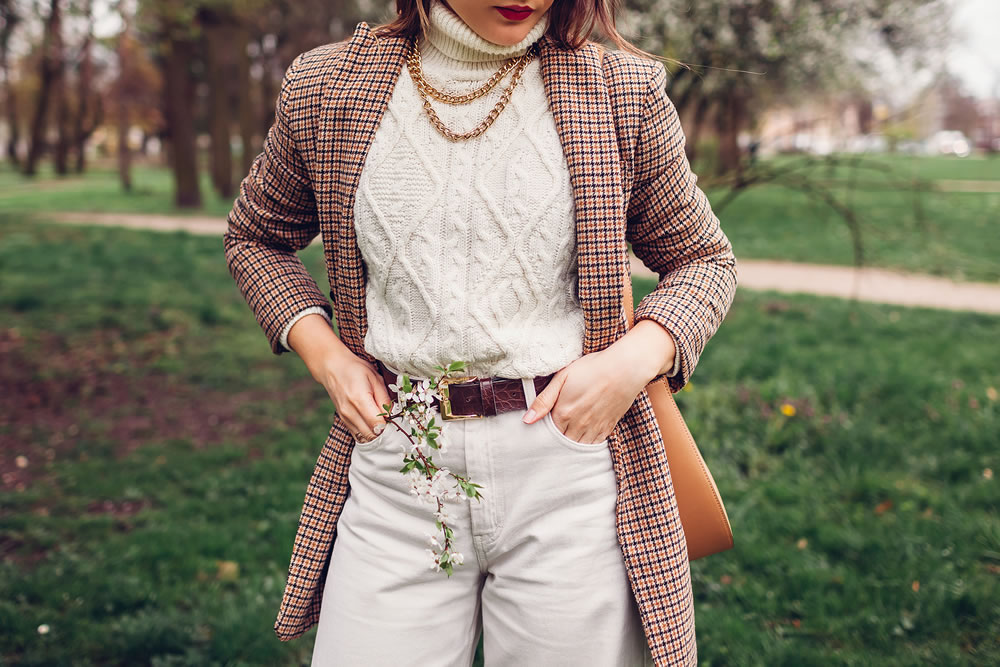 Outdoor close up of young woman wearing white sweater and pants with brown blazer holding purse