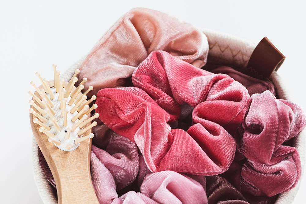 Pink velvet scrunchies and small wooden bamboo hair brush in basket for accessories on white background