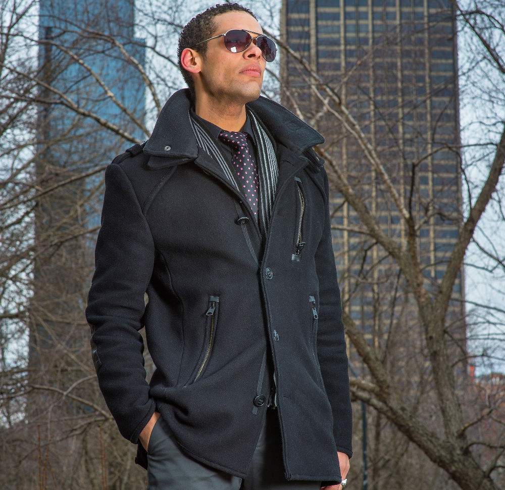 Dressing in a black wool pea coat and a tie, wearing sunglasses a young guy is standing outside and looking forward