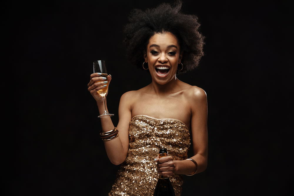 Excited african american woman in party dress drinking champagne isolated over black background