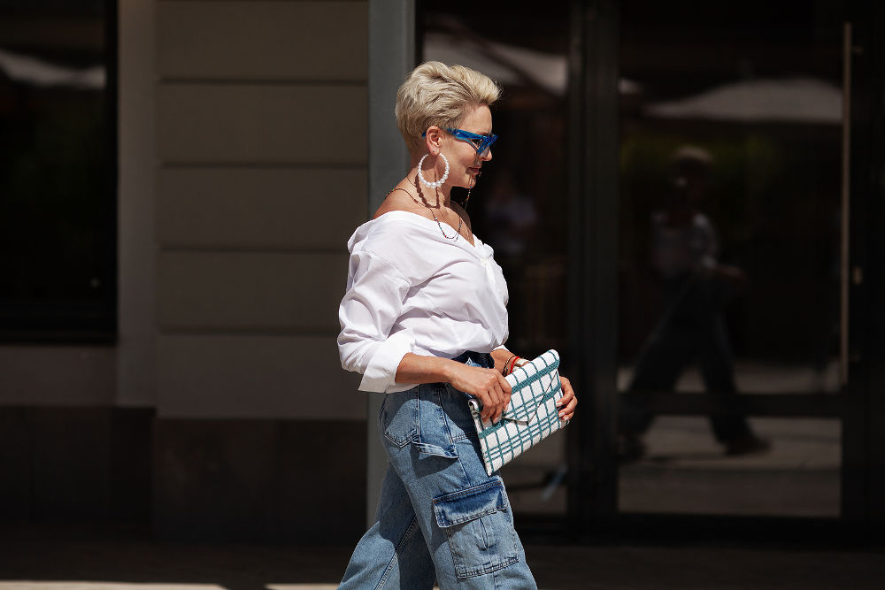 Sophisticated woman with short hair in white shirt and cargo jeans pants, fashionable accessories earrings, stylish glasses and clutch walking outdoors in city