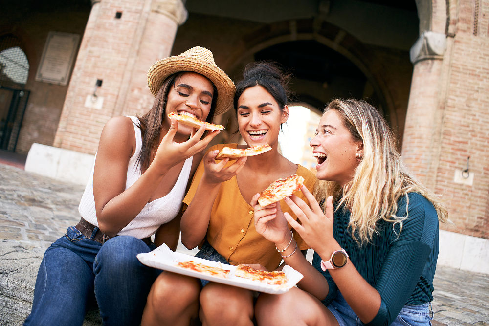 Three funny multiracial women eating pizza outdoors in the street.
