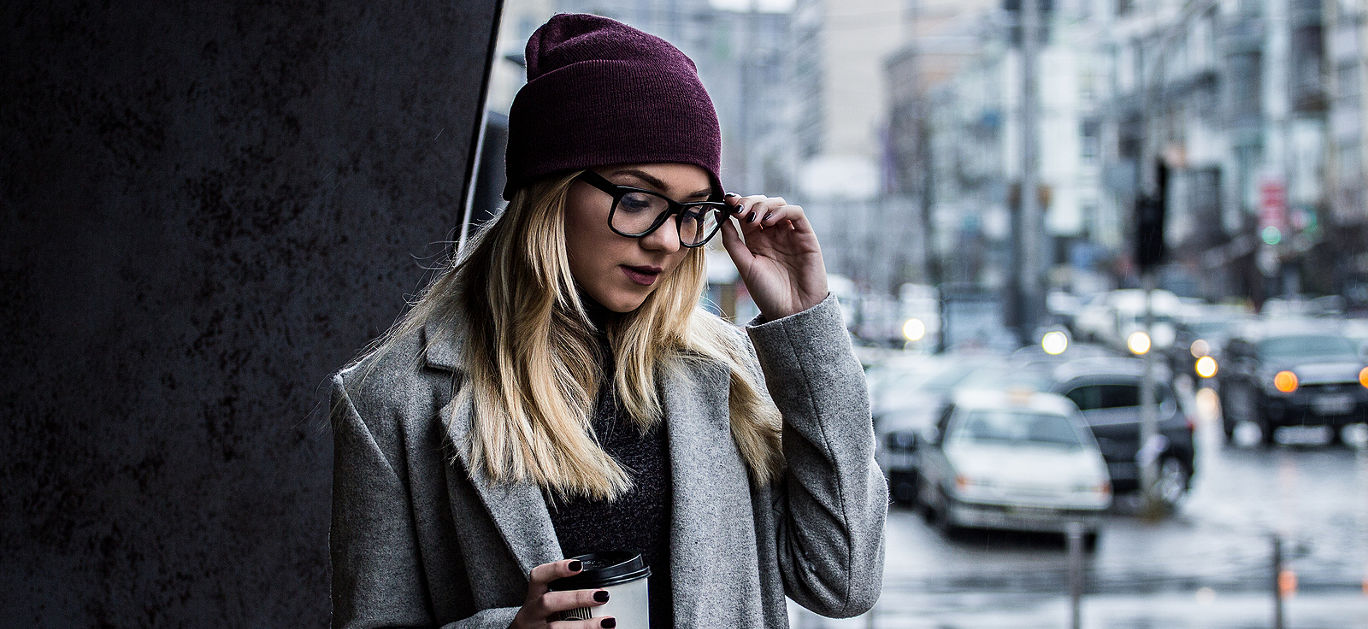 Woman with perfect city style. Portrait of stylish hipster girl in coat holding glasses and looking down while standing outdoors. Young woman in eyeglasses drinking coffee. Urban fashion