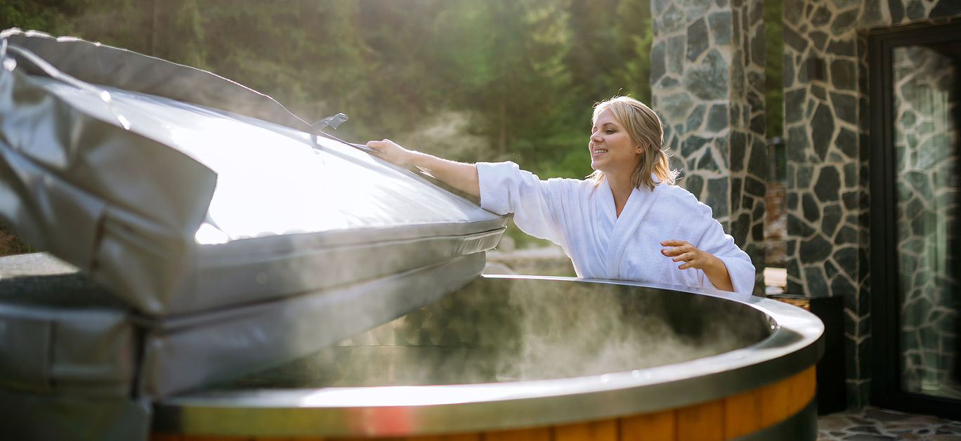 Woman in bathrobe opening lid of hot tub, checking temperature, ready for home spa procedure in hot tub outdoors. Wellness, body care, hygiene concept.